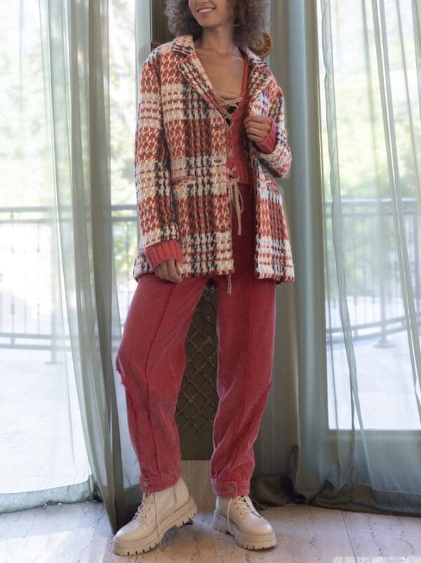 A woman in red pants and plaid jacket standing next to window.